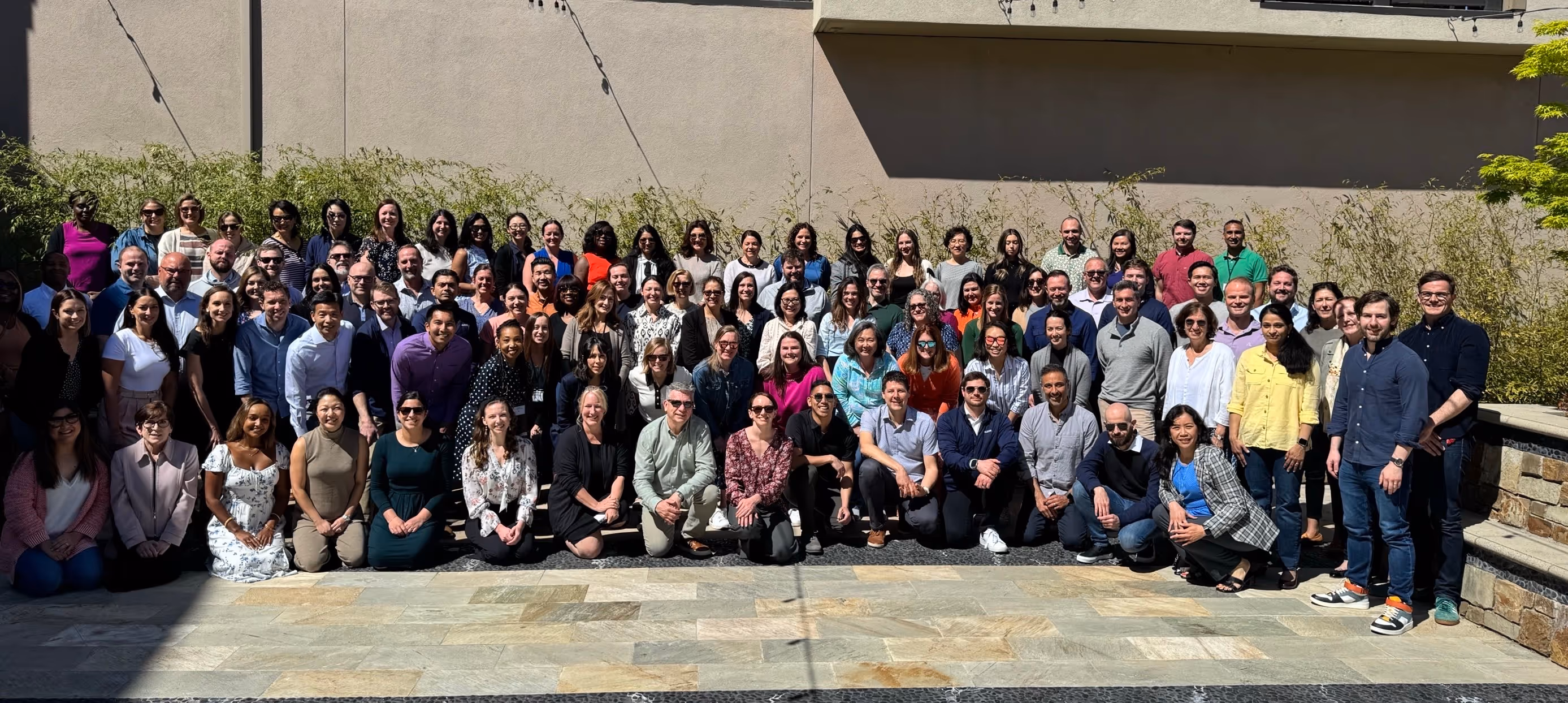 A large group photo of employees gathered outdoors in front of a neutral wall and greenery. The team is smiling in bright sunlight, showcasing diversity and unity in a professional setting.