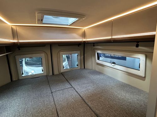 Innenansicht eines Wohnmobils mit gemütlichem Bett, Deckenbeleuchtung und kleinen Fenstern im Hintergrund.