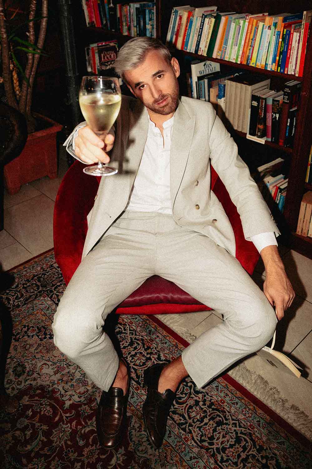 Der Musiker Leo Poldau sitzt mit Weinglas in der Hand auf einem roten Stuhl in einer Bücherei.