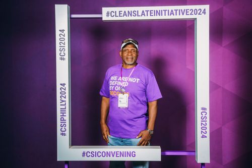A man wearing a purple shirt and a badge stands in front of a purple background.
