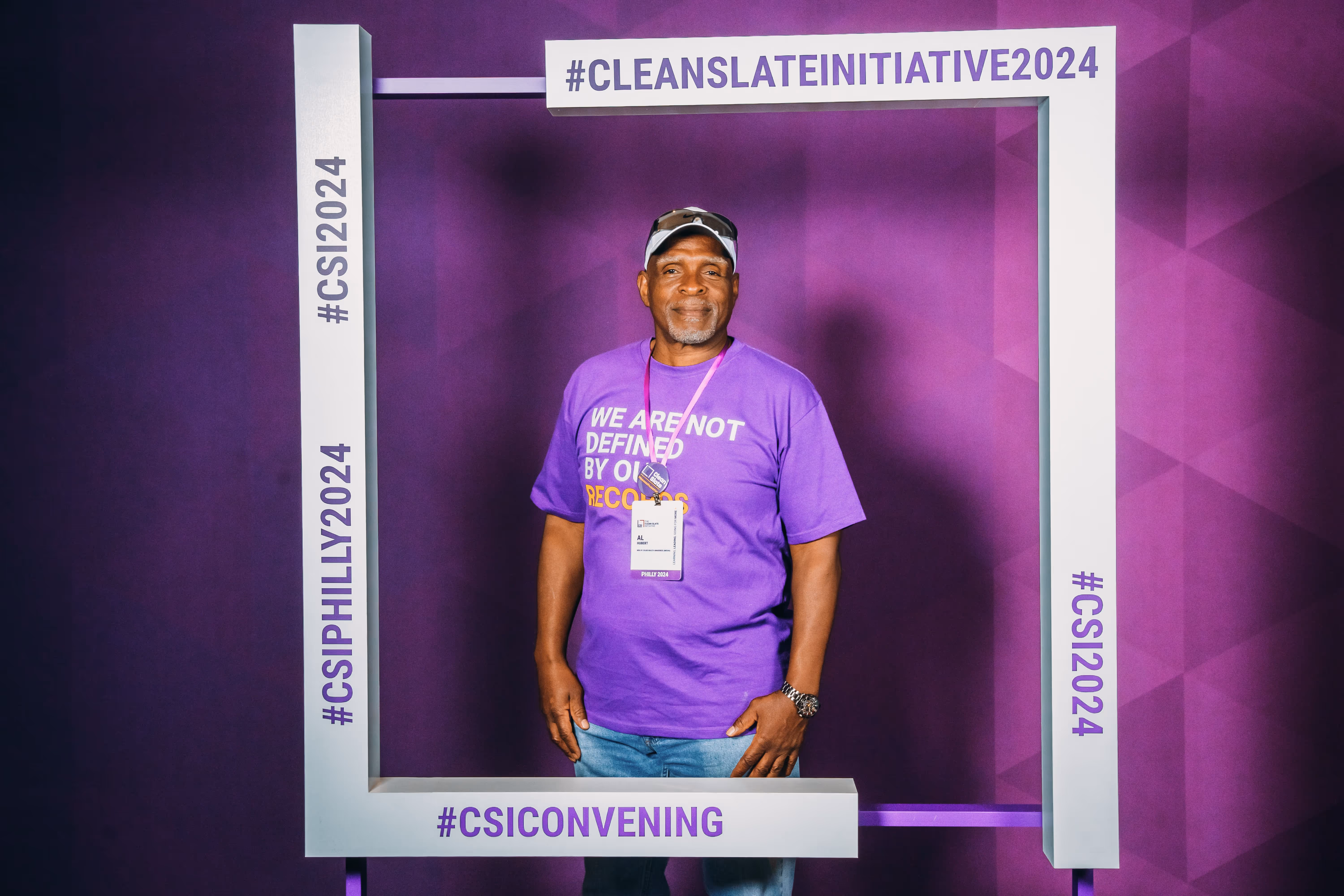 A man wearing a purple shirt and a badge stands in front of a purple background.