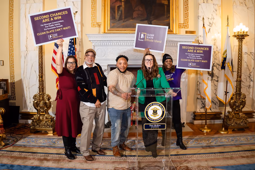 A diverse group of advocates stands behind a podium holding signs in support of Clean Slate in Rhode Island
