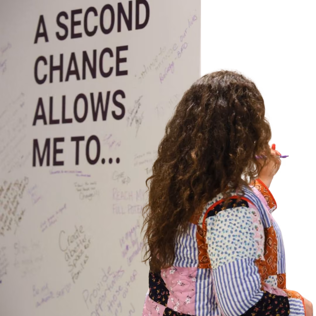 A woman stands in front of a wall, adding her own answer to the prompt "A Second Chance Allows Me To...", among other hand-written answers to the same prompt.