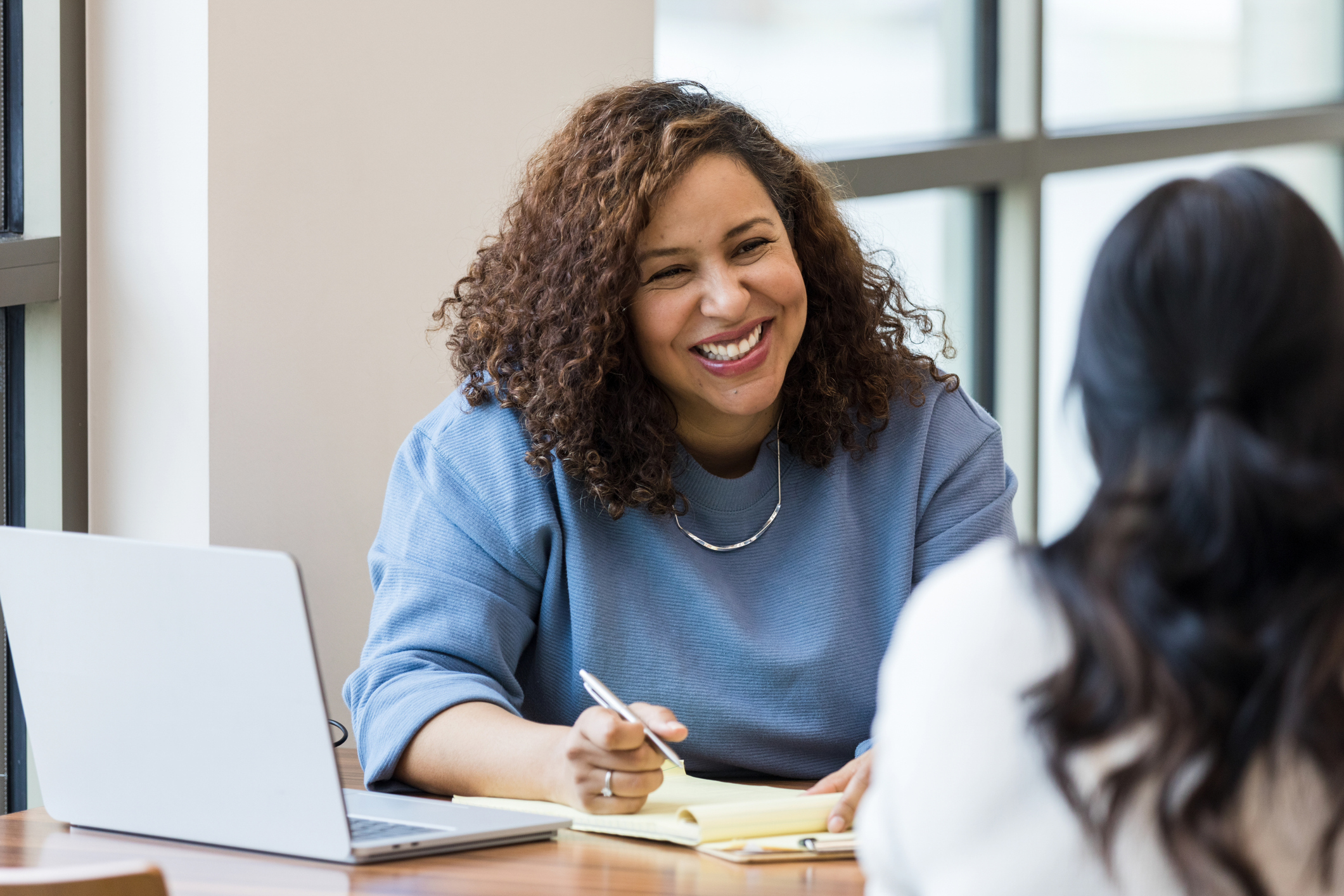 Smiling woman with curly hair writing on a notepad during a meeting with another person.