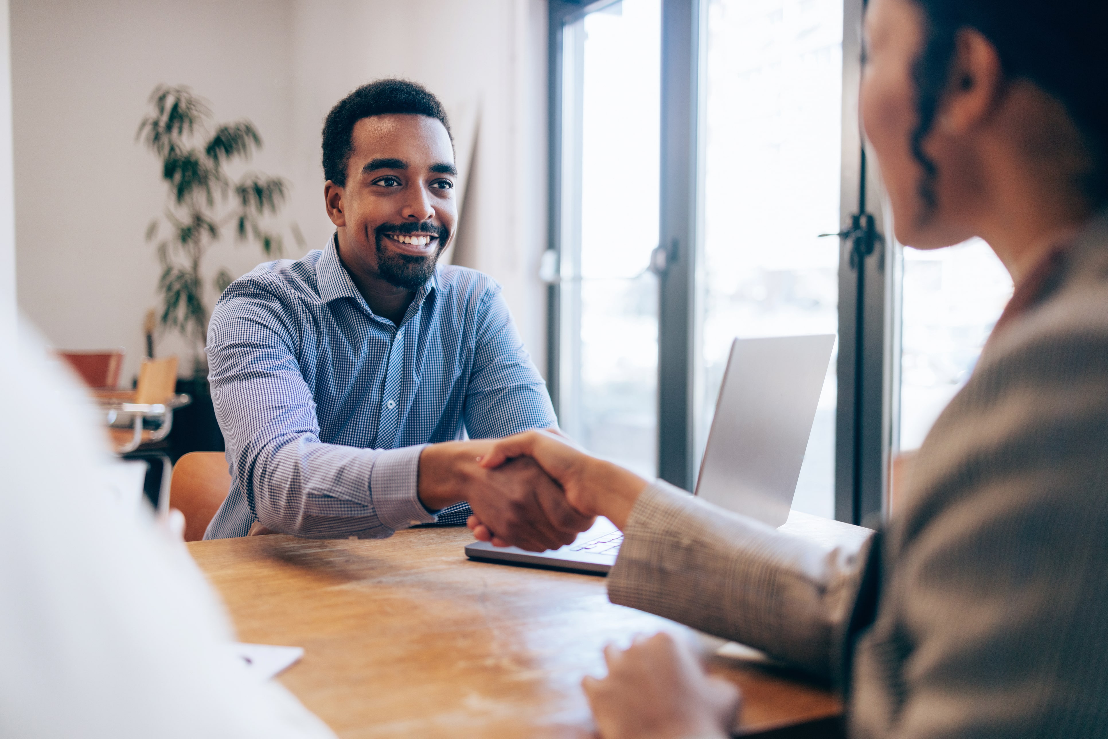 Smiling man in blue checkered shirt shaking hands with a woman during a meeting at a wooden table with a laptop.