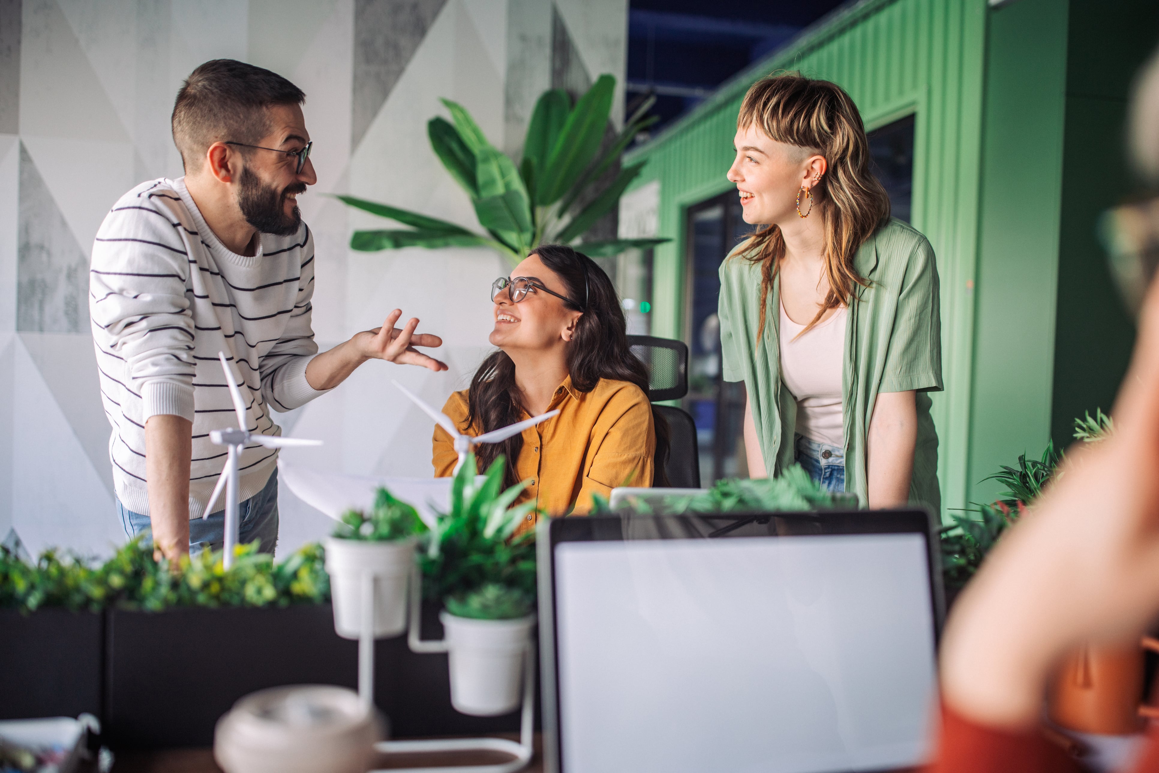 Three coworkers smiling and discussing in a modern office with green plants and a laptop nearby.