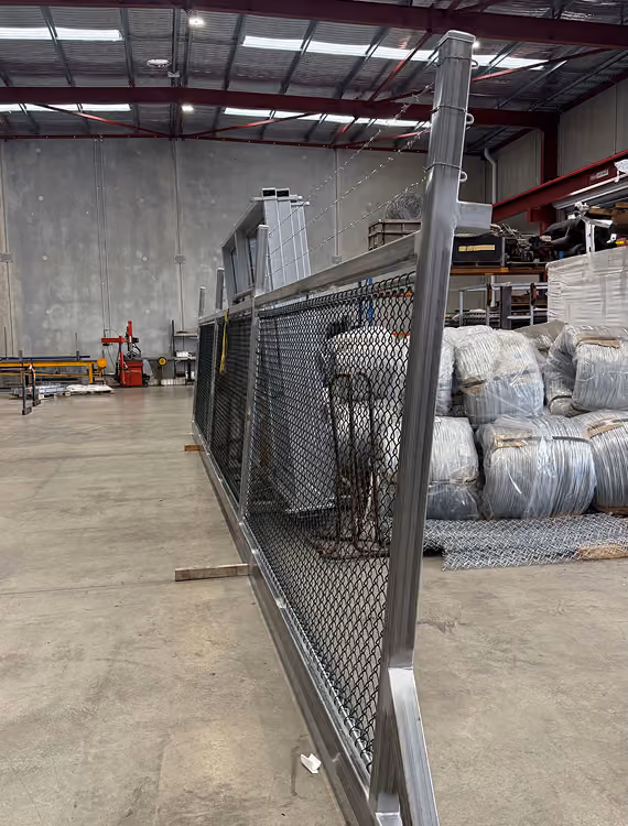 Long chain-link fence panel with metal frame standing vertically inside a warehouse with a concrete floor and high ceiling, with large wrapped pallets and industrial equipment in the background.