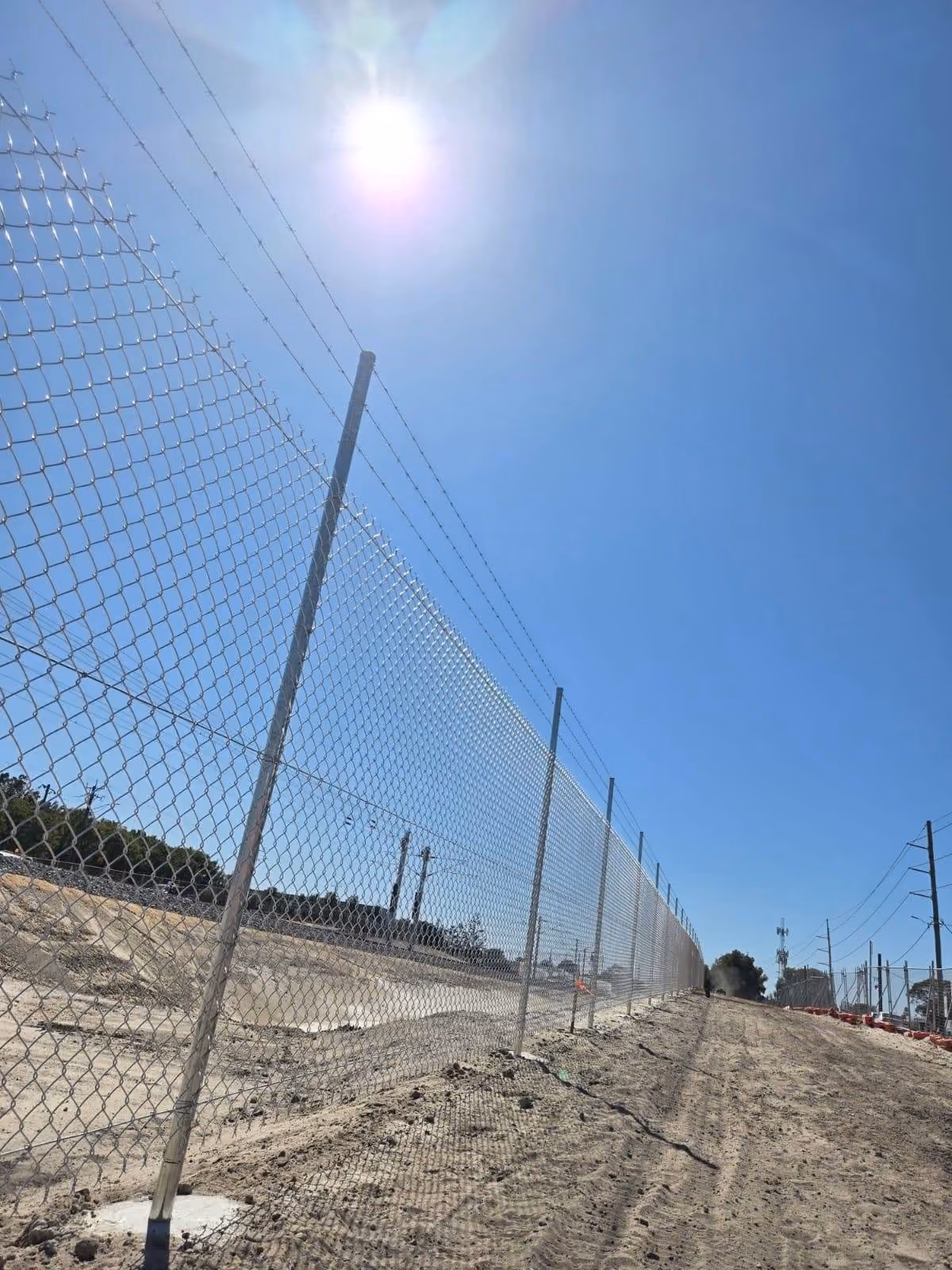 Chain-link fence with barbed wire on top running diagonally alongside a dirt path under a bright sunny sky.
