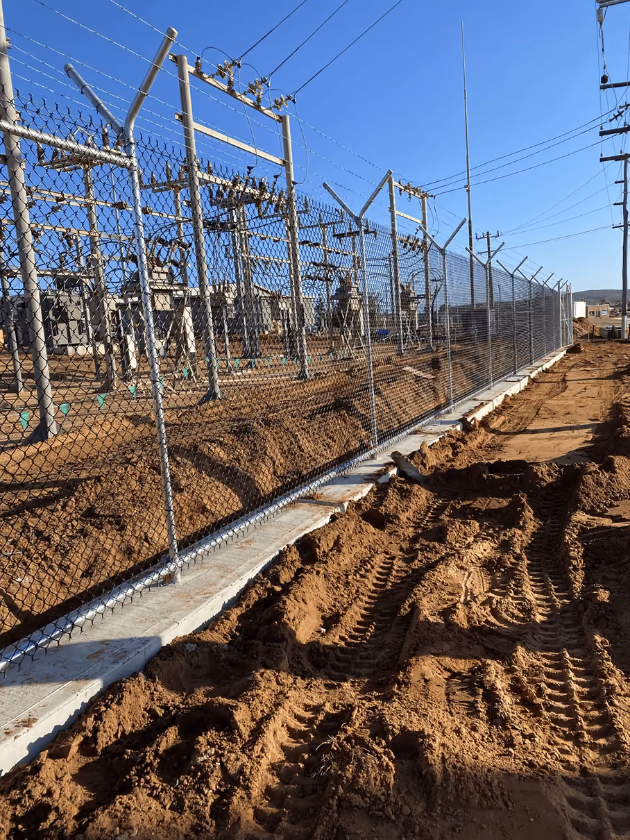 Chain-link fence topped with barbed wire enclosing an electrical substation with transformers and power lines under a clear blue sky, with dirt and tire tracks in the foreground.
