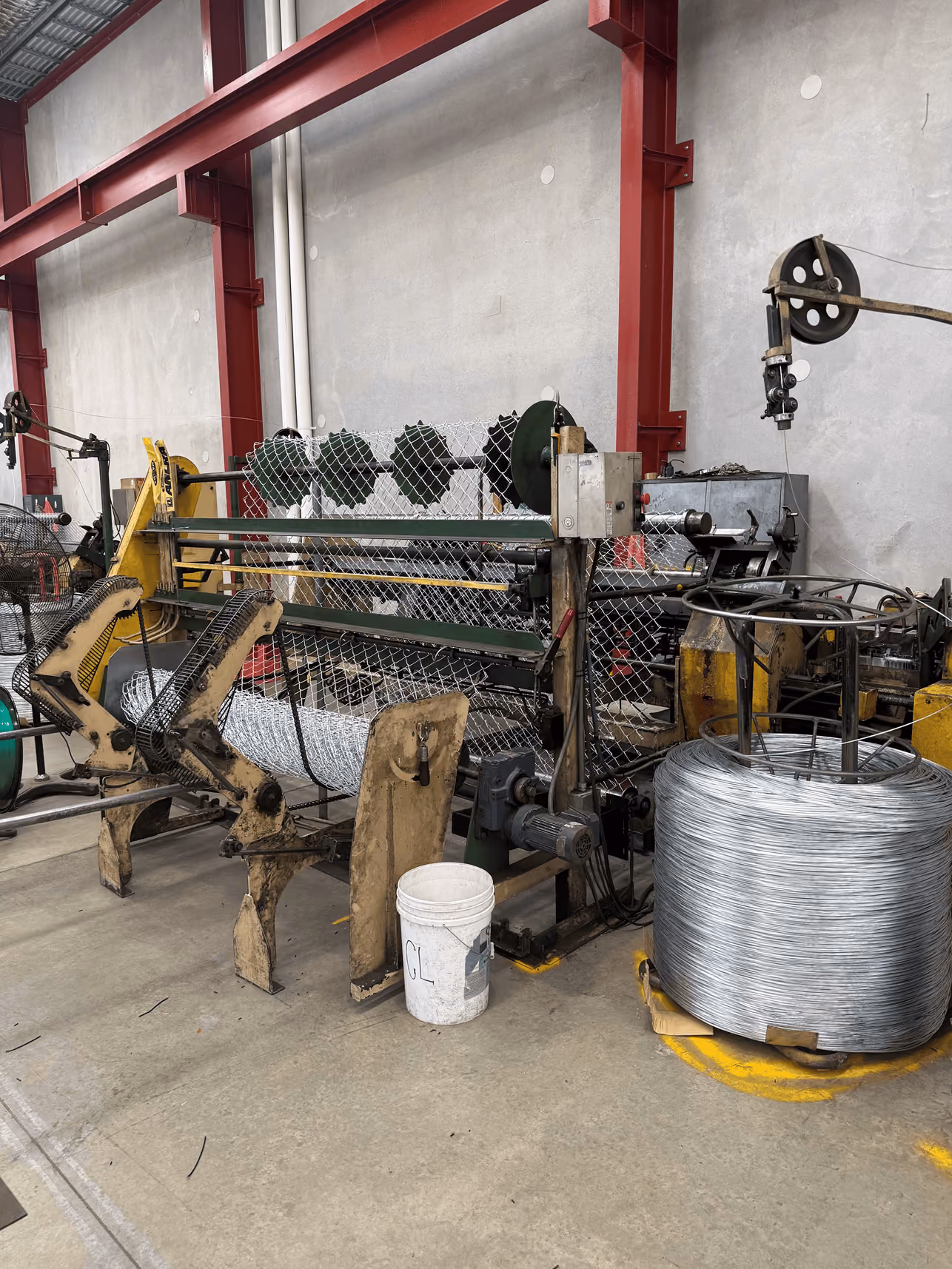 Industrial wire mesh manufacturing machine in a factory with a large coil of metal wire on the floor next to it, red structural beams, and a white plastic bucket labeled 'CL' nearby.