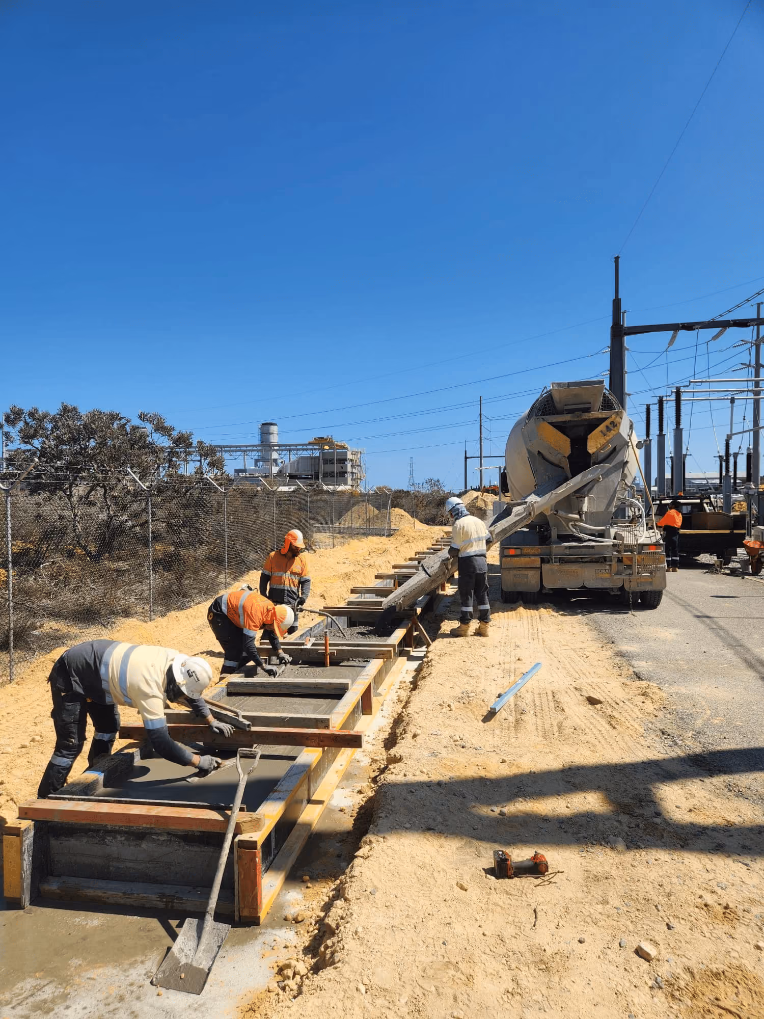 Construction workers in safety gear pouring and smoothing concrete in wooden molds along a sandy path next to a cement mixer truck under clear blue sky.