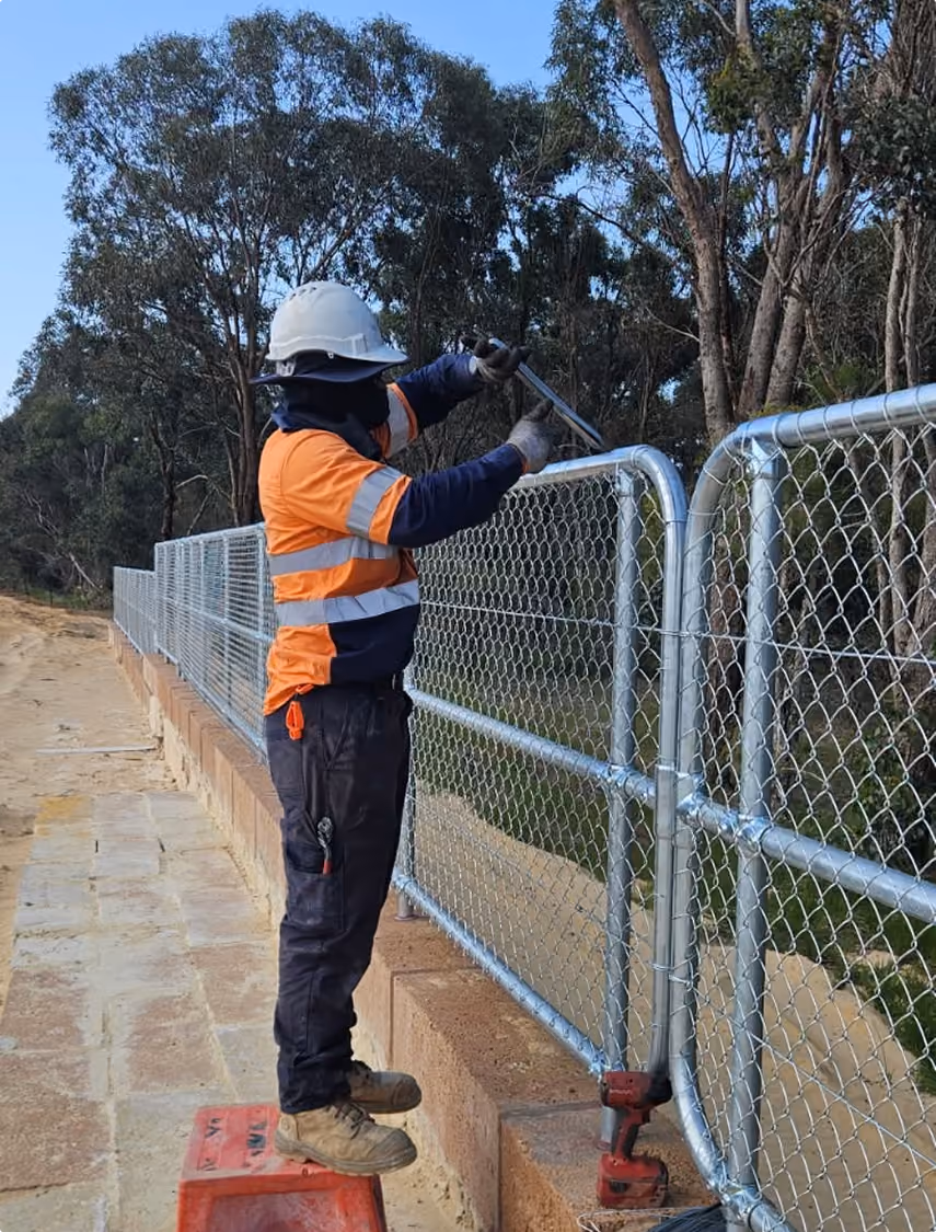 Construction worker wearing a white hard hat, orange high-visibility shirt, and dark pants, standing on a red crate while using a wrench to adjust a metal chain-link fence outdoors, with trees and clear sky in the background.