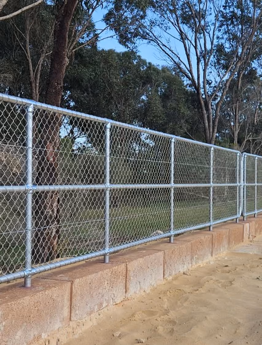 Chain link fence mounted on a low concrete wall beside a sandy ground with trees and blue sky in the background.