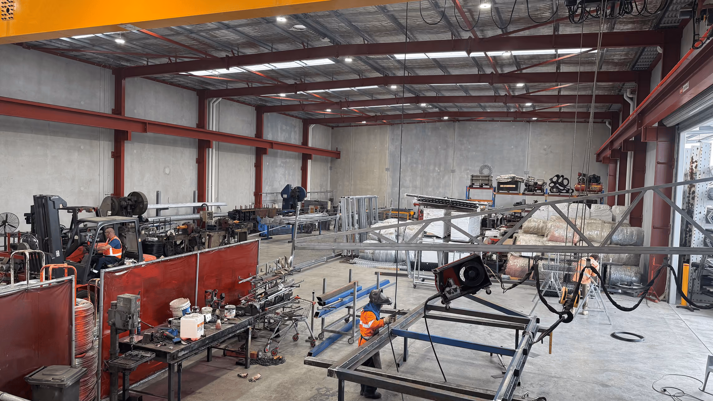 Interior of a large industrial workshop with workers wearing orange safety vests operating machinery and assembling metal structures, surrounded by tools, equipment, and industrial materials.