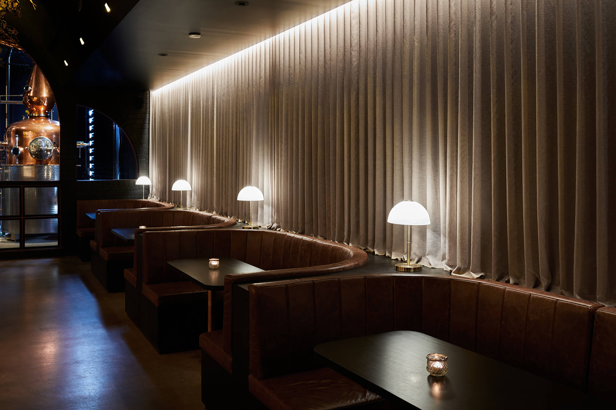 Curved Cognac Leather Booths Dark Tables Brass Opal Lamps Sheer Linen Drapes Linear Cove Lighting in Naught Distilling. Photo by Graham Denholm.