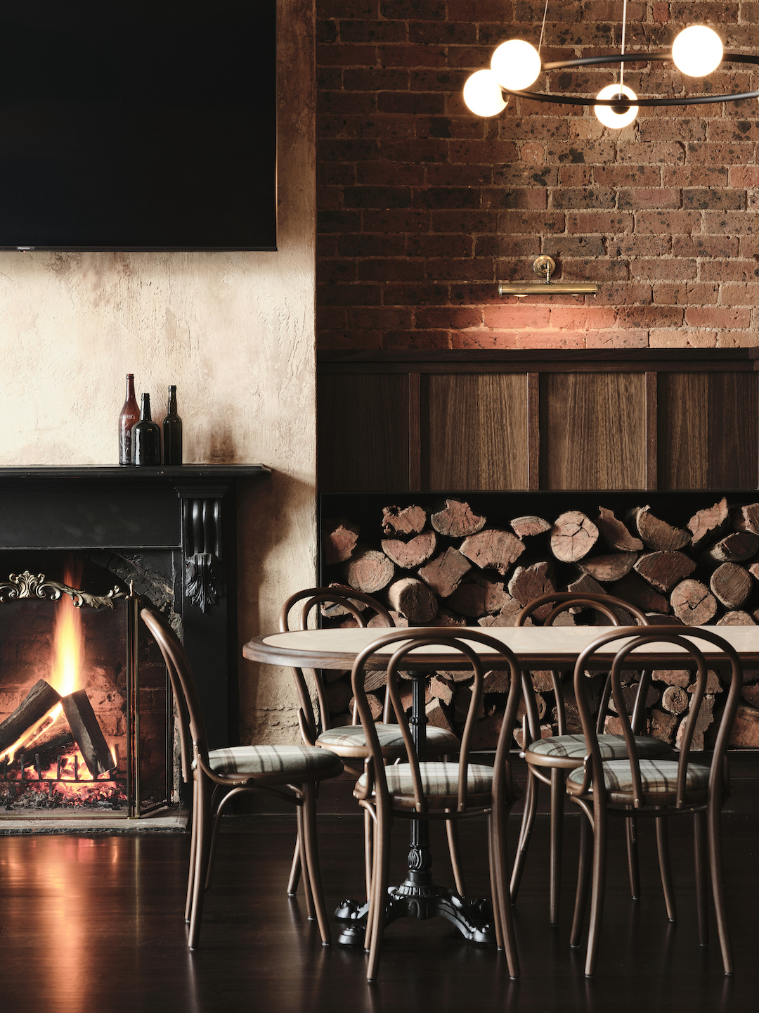 Historic Fireplace with Ornate Black Mantelpiece Exposed Brick Wall Stacked Log Storage Bentwood Dining Chairs and Warm Lighting in The Bay Hotel. Photo by James Geer.