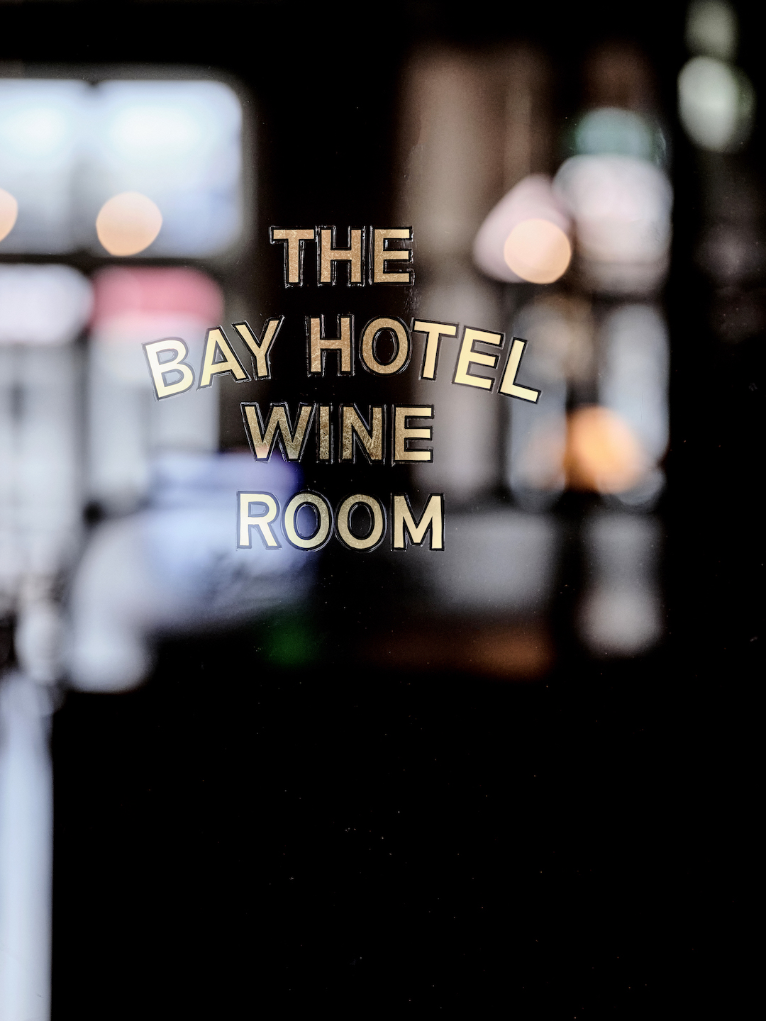 Gold Leaf Decal Typography on Glass Door Dark Moody Backdrop with Soft Bokeh Highlights Traditional Signage Aesthetics in The Bay Hotel. Photo by James Geer.
