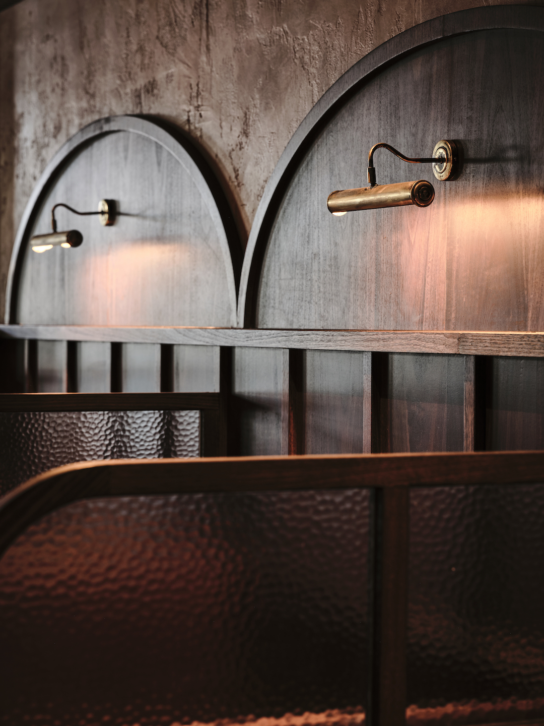 Aged Brass Sconces on Arched Walnut Panels Hammered Glass Partitions Textured Rendered Walls Warm Ambient Glow Heritage Detail in The Bay Hotel. Photo by James Geer.