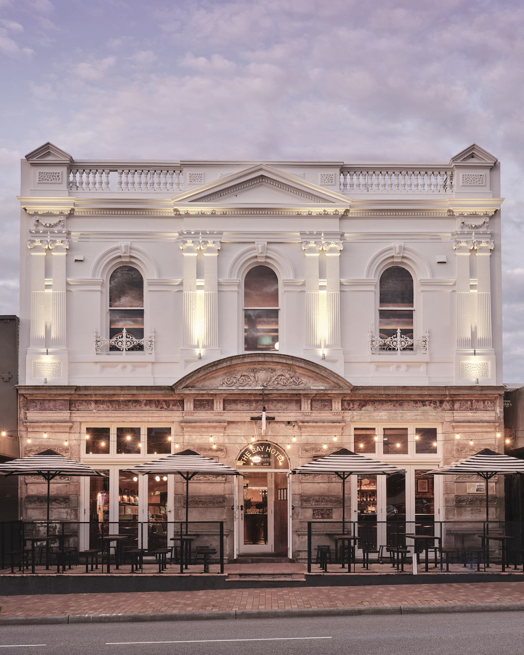 Neoclassical Facade with Corinthian Pilasters Parapet Balustrade Alfresco Dining Striped Umbrellas and Festoon Lighting in The Bay Hotel. Photo by James Geer.