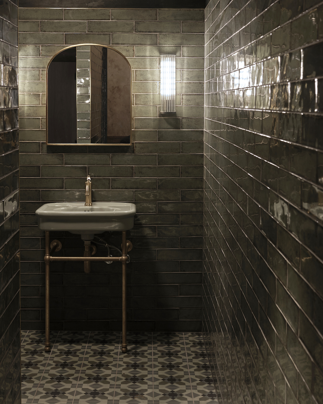Glossy Olive Subway Tiles Arched Brass Mirror Fluted Wall Sconce Pedestal Basin with Industrial Gold Piping and Patterned Floor Tiles in The Bay Hotel. Photo by James Geer.