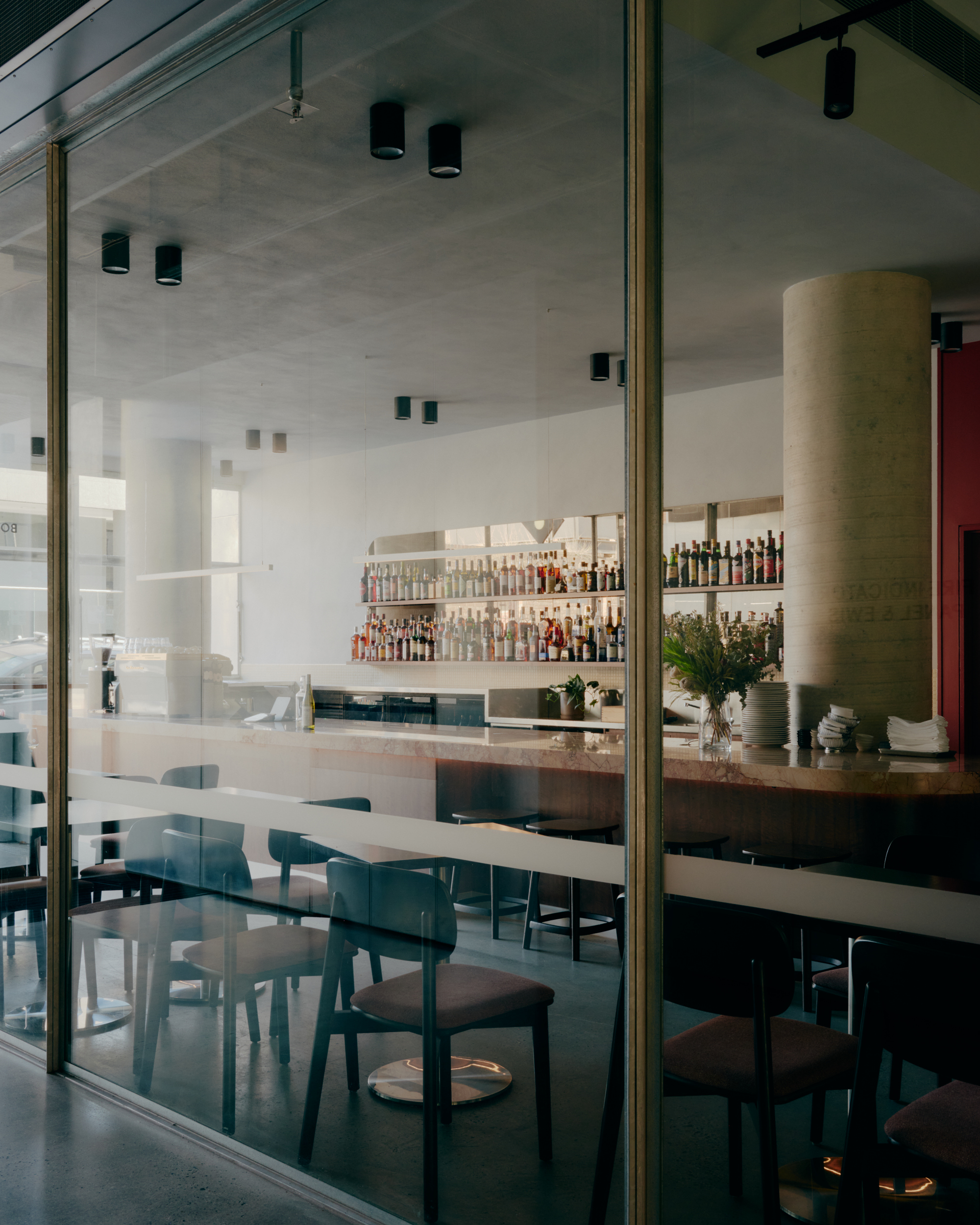 View Through Glazing of Minimalist Bar Area with Rose Marble Counter Extensive Spirits Backbar Concrete Columns and Modern Cafe Seating in The Local Drop. Photo by Pier Carthew and Kevin McDowell.