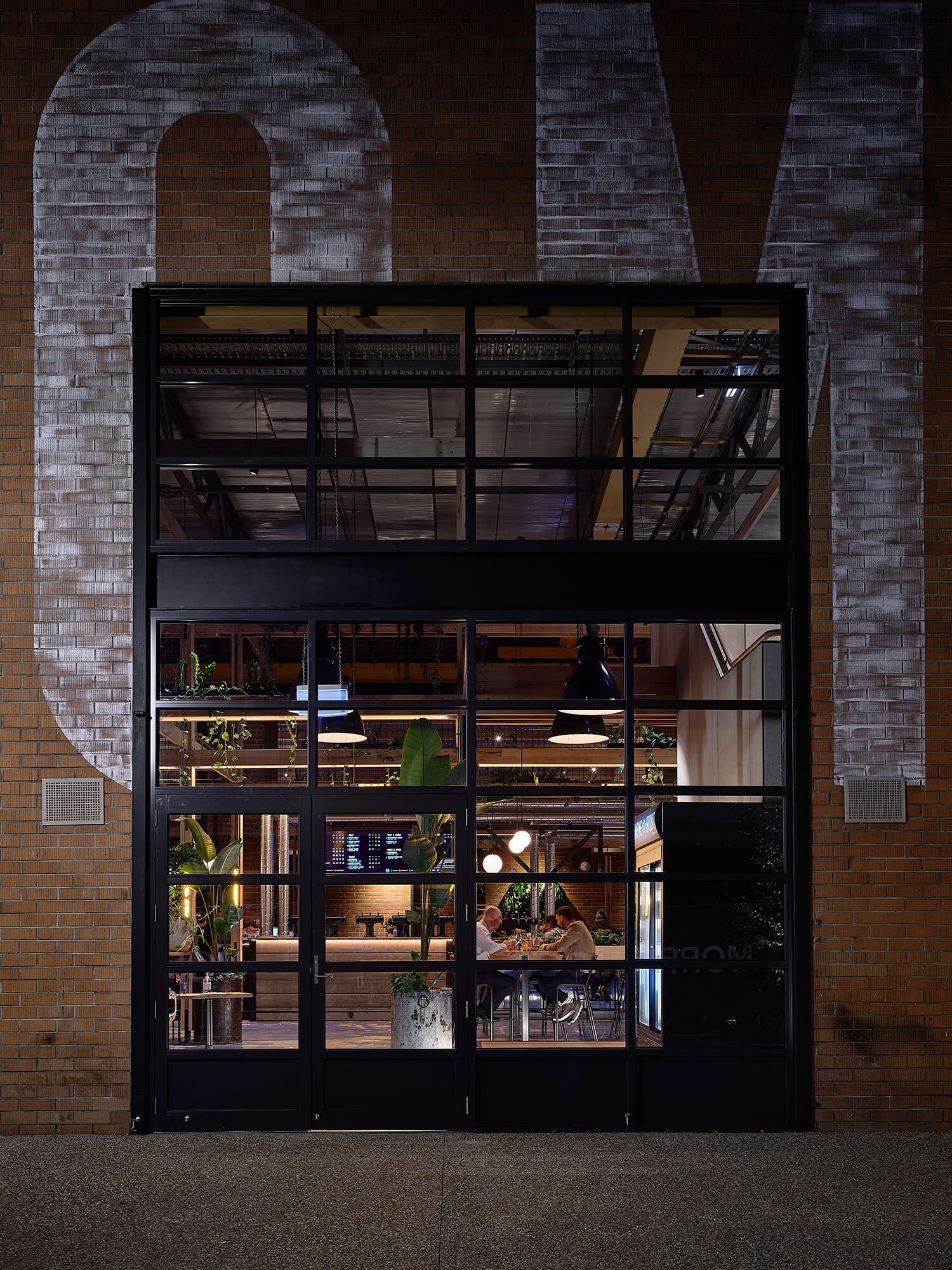 Night View of Industrial Glass Entry Framed by Large Painted Brick Graphics Revealing Warm Interior Lighting and Potted Plants at Stomping Ground Morris Moor. Photo by Derek Swalwell.