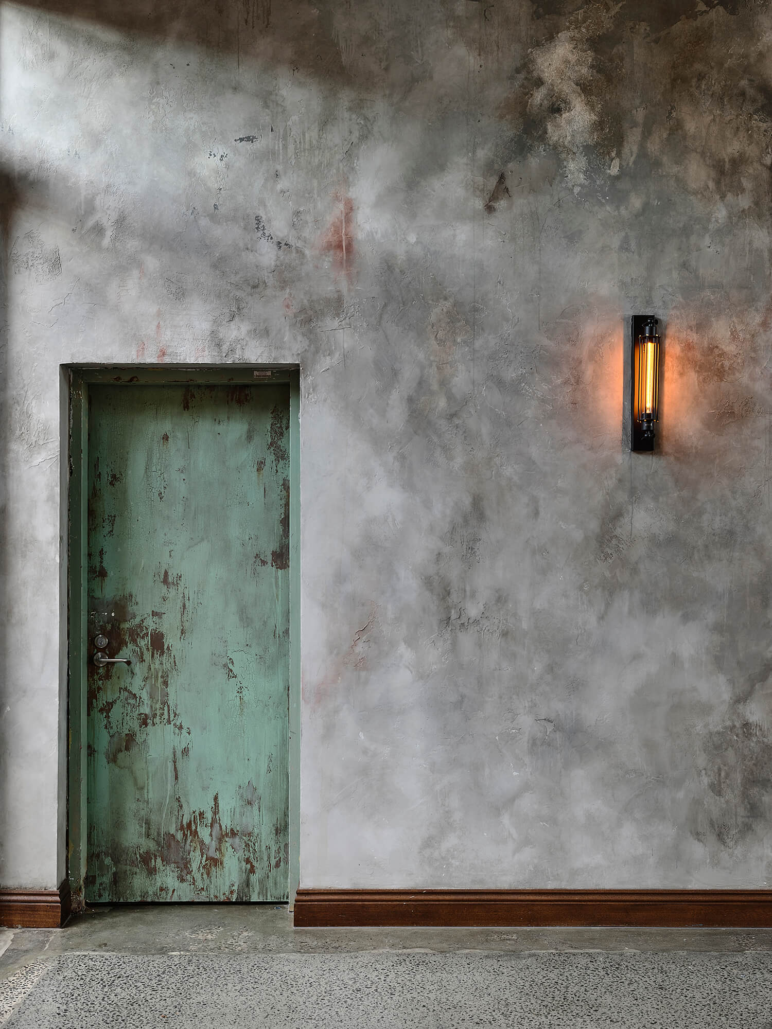 Weathered Green Industrial Door Set in Textured Concrete Wall with Vintage Filament Wall Sconce and Timber Skirting in Stomping Ground Morris Moor. Photo by Derek Swalwell.