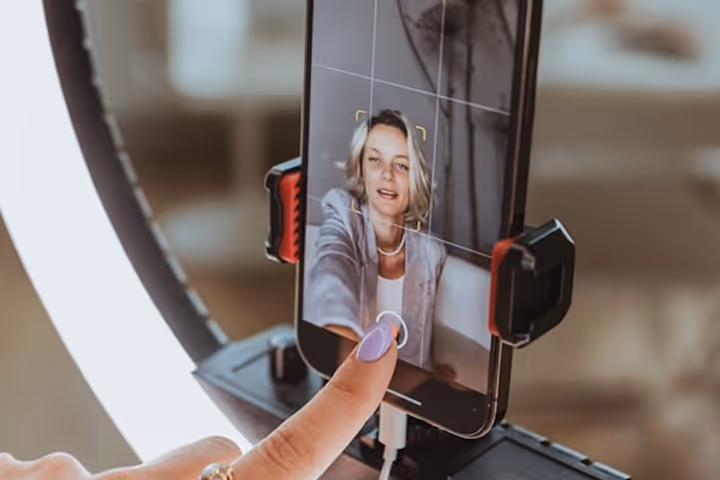 Close-up of a hand with painted nails pressing the capture button on a smartphone screen showing a woman in a video recording setup with ring light.