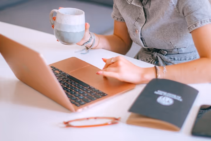 Person holding a white and blue ceramic mug while working on a rose gold laptop at a white desk with glasses and a notebook nearby.