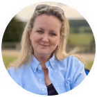 Smiling woman with blonde hair wearing a light blue shirt outdoors with a blurred green background.