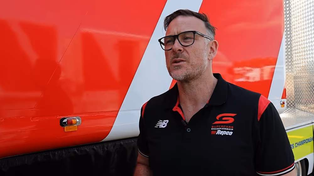 Man with glasses speaking in front of a red and white vehicle, wearing a black polo shirt with Supercars Championship and Repco logos.