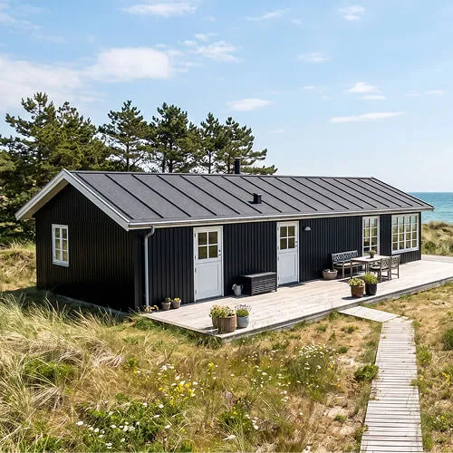 Sort sommerhus ved kysten med træterrasse og havudsigt omgivet af græs og klitter.