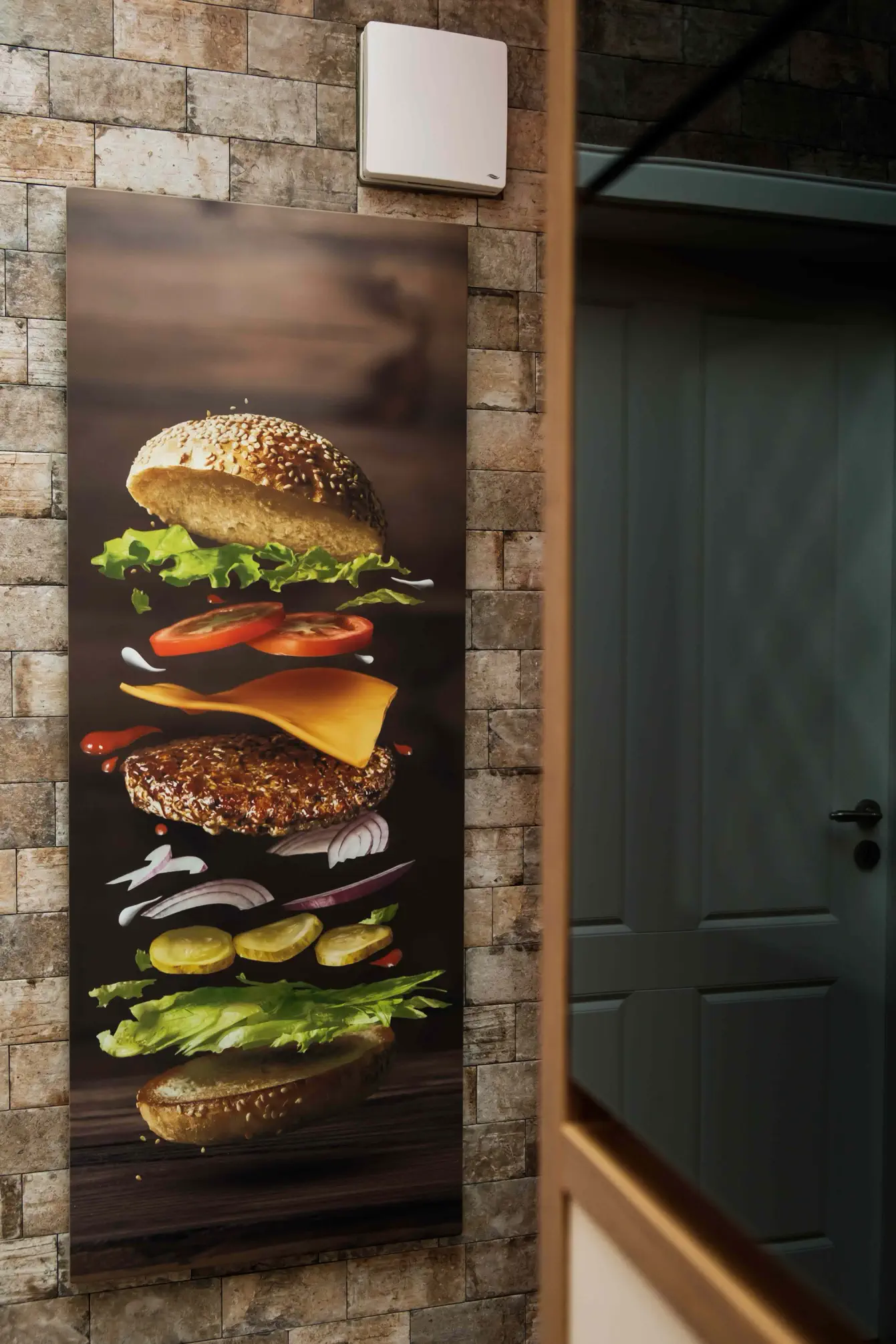Aflangt infrarødt varmepanel fra Redwell med trykt motiv af en burger i flere lag. Billedvarmen er monteret på en murstensvæg ved siden af en indgang og fungerer som dekorativt skilt, f.eks. i en burgerbar eller restaurant.