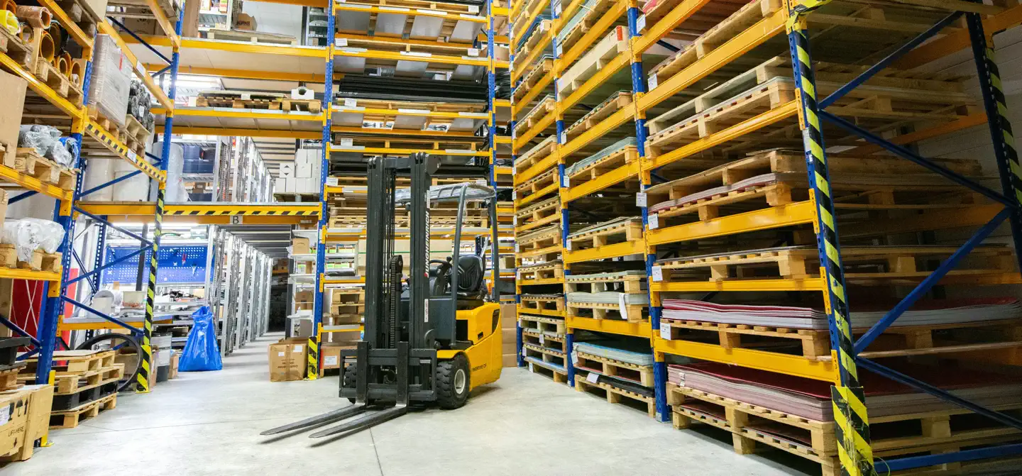 Kraftige Redwell Powerline infrarøde varmepaneler monteret i loftet på et stort, lyst lager med gule og blå palleløftere og en gaffeltruck. Viser effektiv industriopvarmning i rum med højt til loftet.