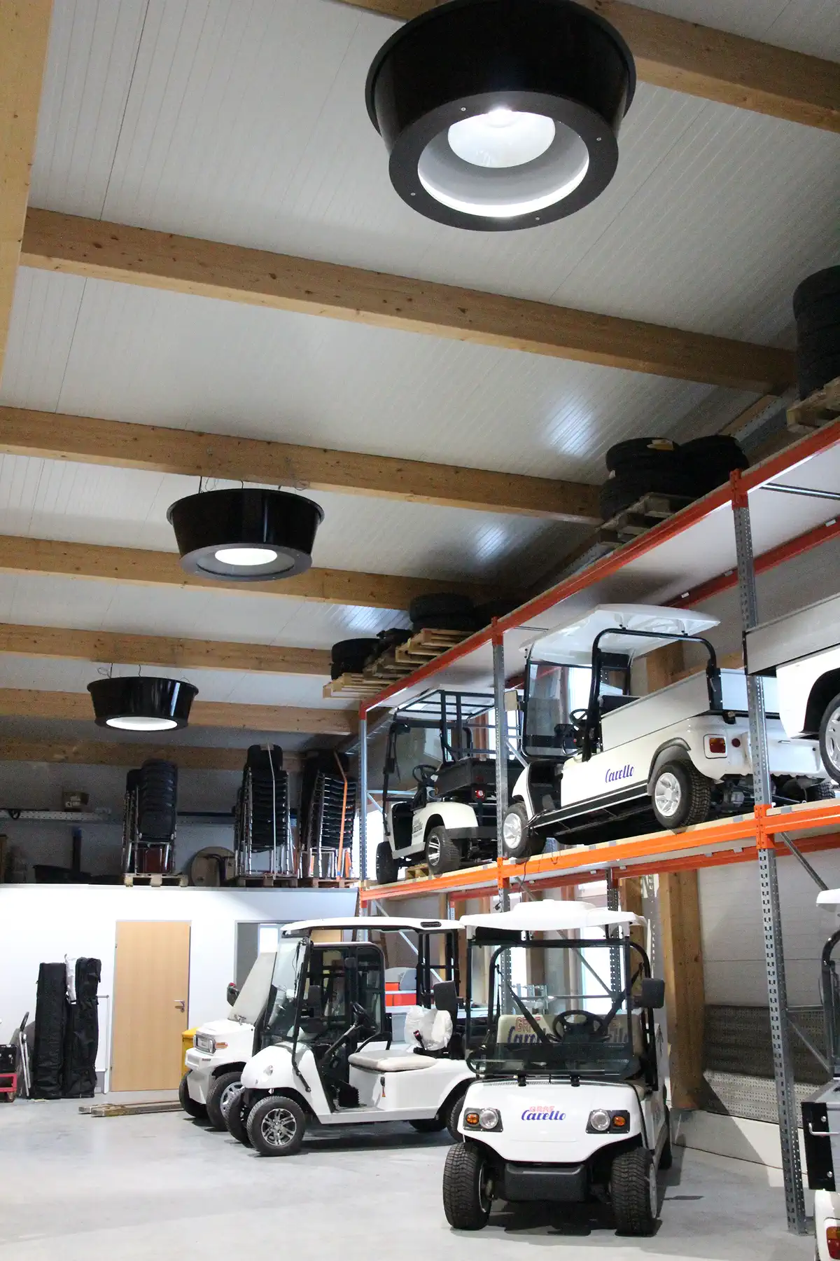 Flere sorte Redwell 2-i-1 loftlamper monteret på træbjælkerne i loftet af en stor lagerhal med parkerede golfvogne. Lamperne kombinerer effektiv infrarød varme og belysning i én energibesparende løsning til store rum.