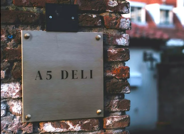 Kvadratisk Redwell infrarødt varmepanel med trykt firmalogo 'A5 DELI', monteret som opvarmet skilt på en rustik murstensvæg.
