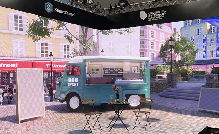 A scene from within a virtual set design based on the squares of Montmartre, Paris. A Cirtoen HY coffee van in vintage BBC colours stands on cobbled paving, and is surrounded by trees and Parisian Apartments. The whole scene is displayed inside large format LED screens that comprise the XR stage at Portsmouth Universities CCIXR