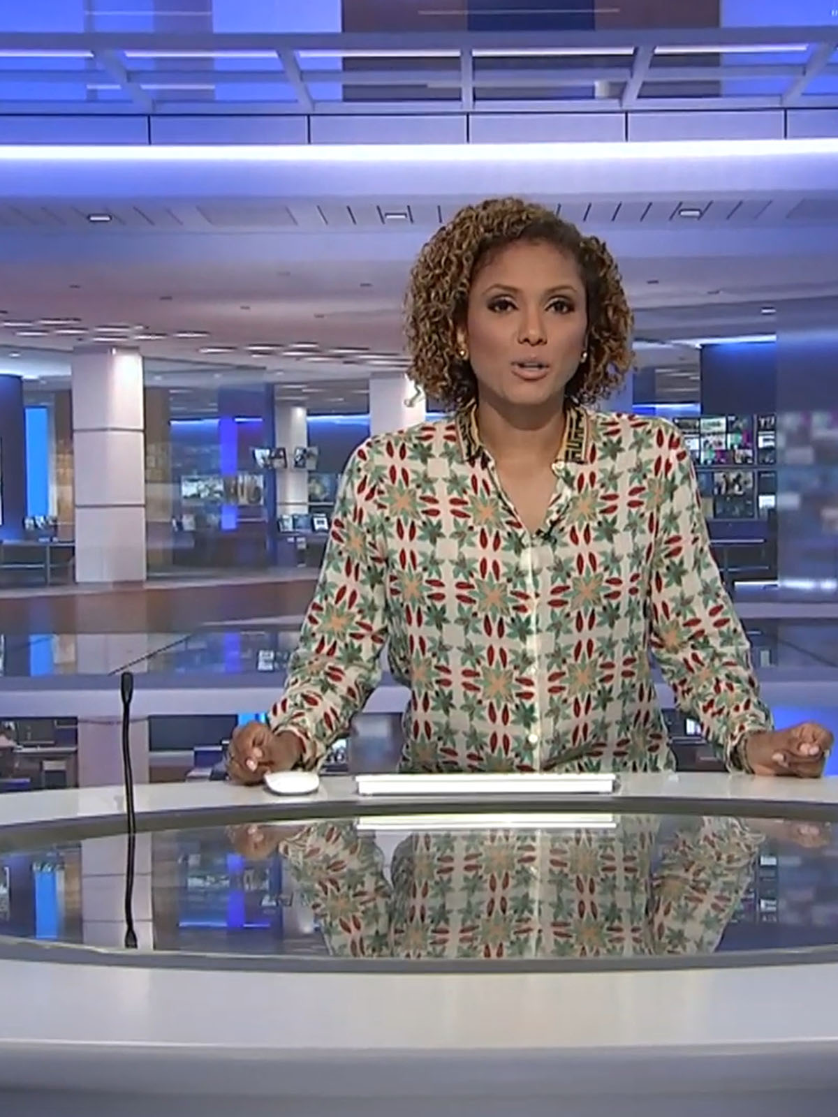 Female news anchor with curly hair at a desk in a modern newsroom with screens behind her.