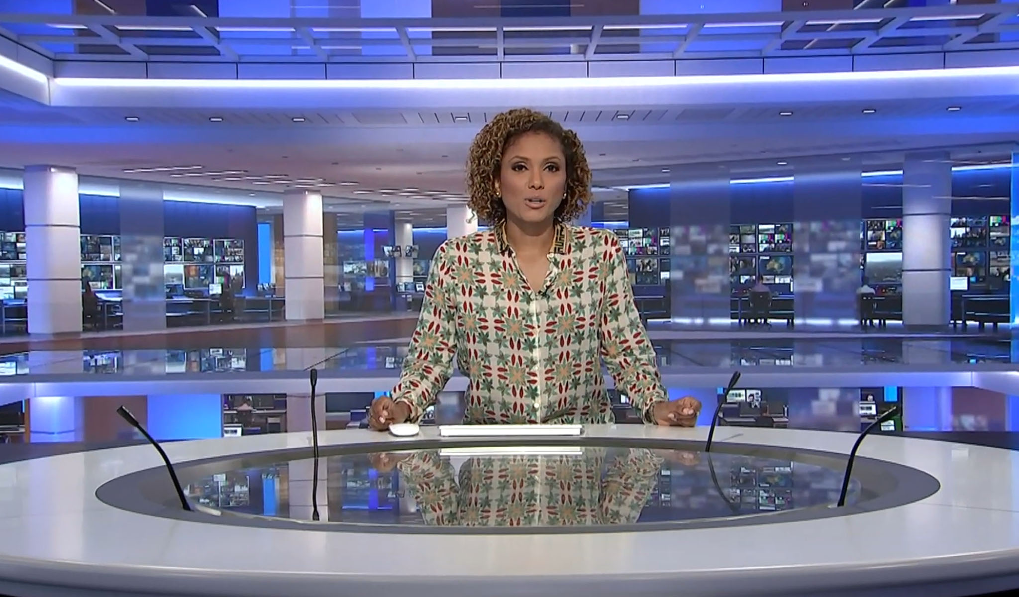 Female news anchor in patterned blouse standing behind a modern news desk with screens in background.