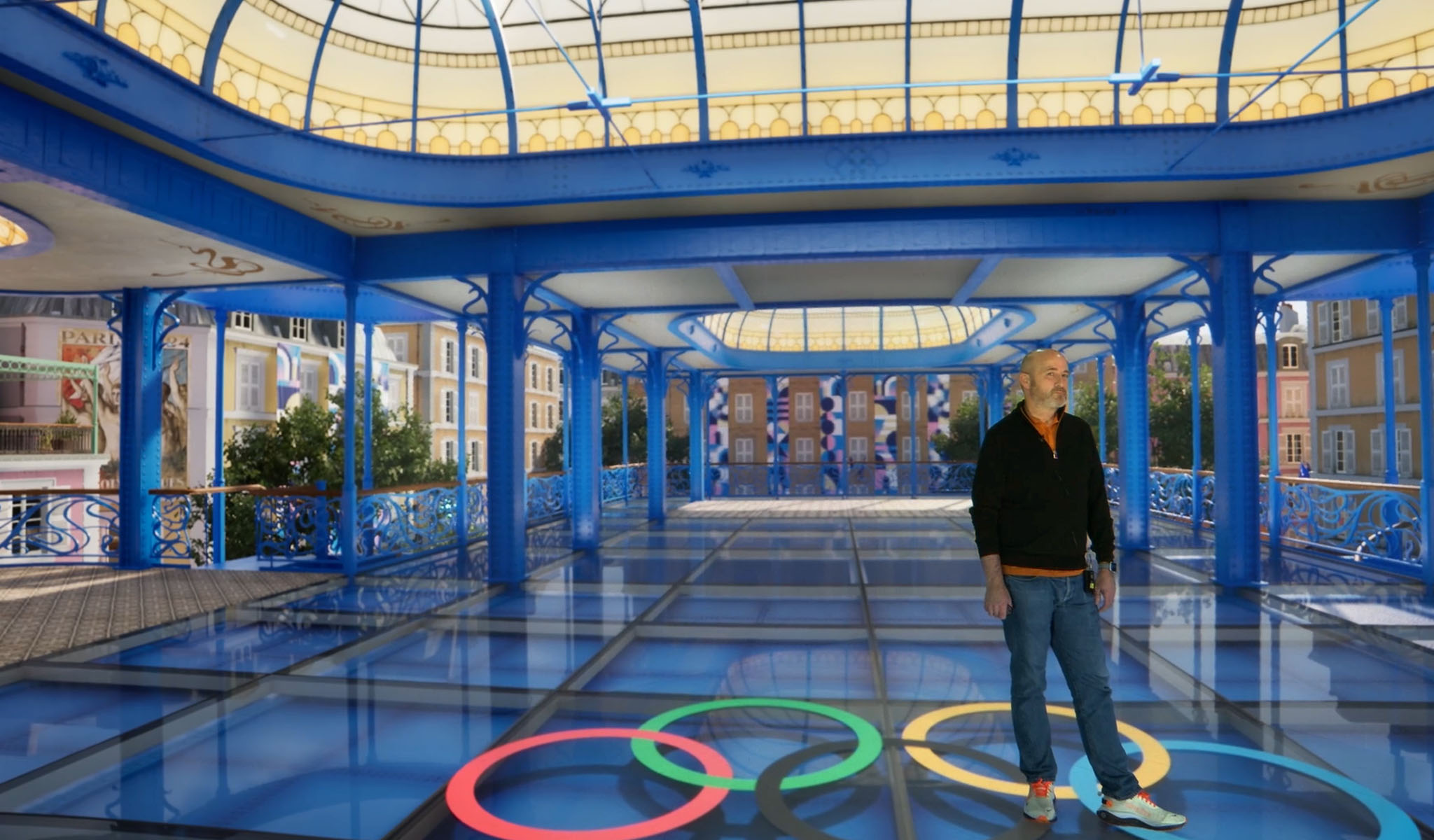 Man stands on glass floor with Olympic rings under blue metal and glass canopy in urban setting.