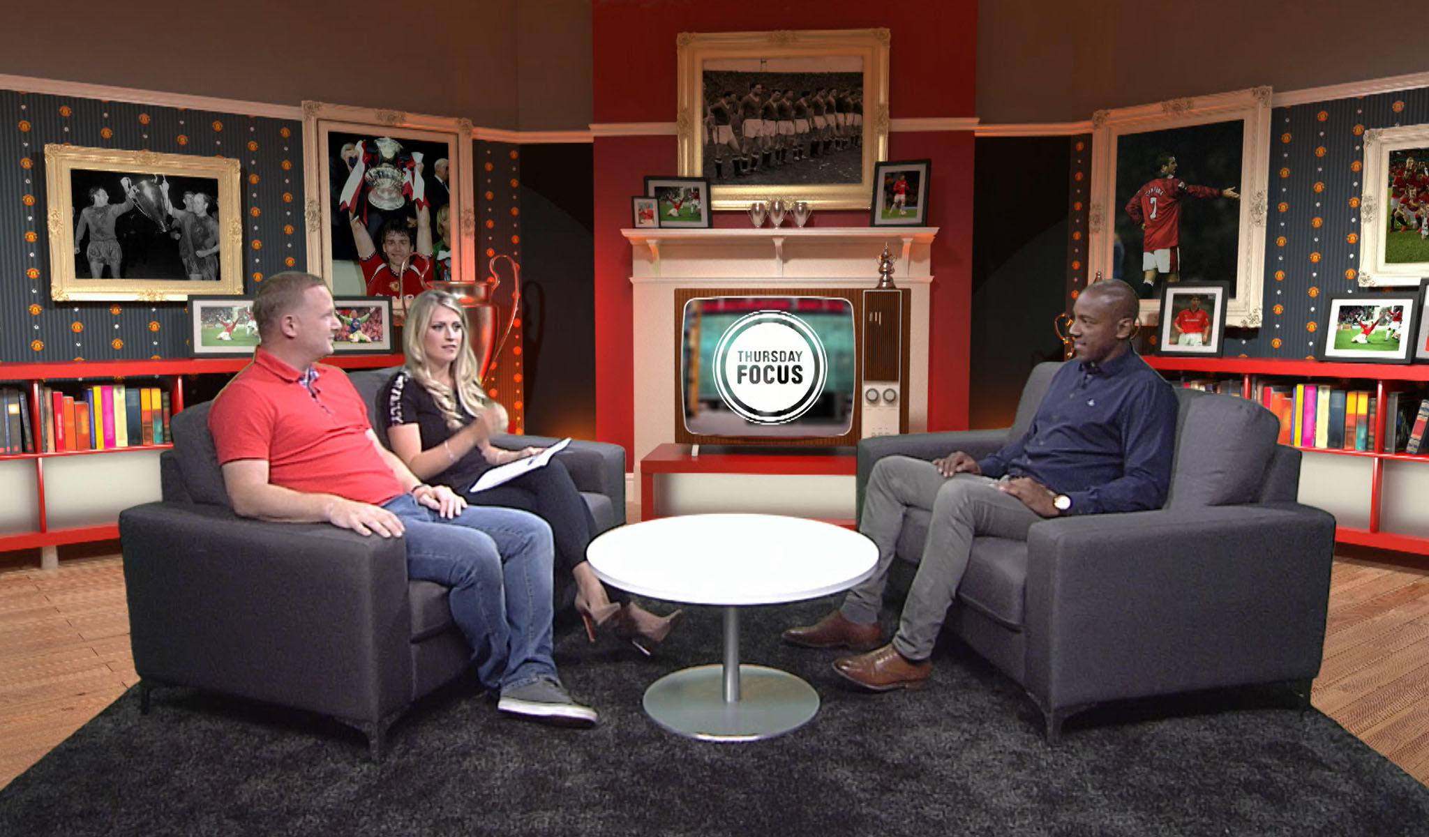 Three hosts seated on gray chairs in a studio with sports photos and a retro TV displaying Thursday Focus.