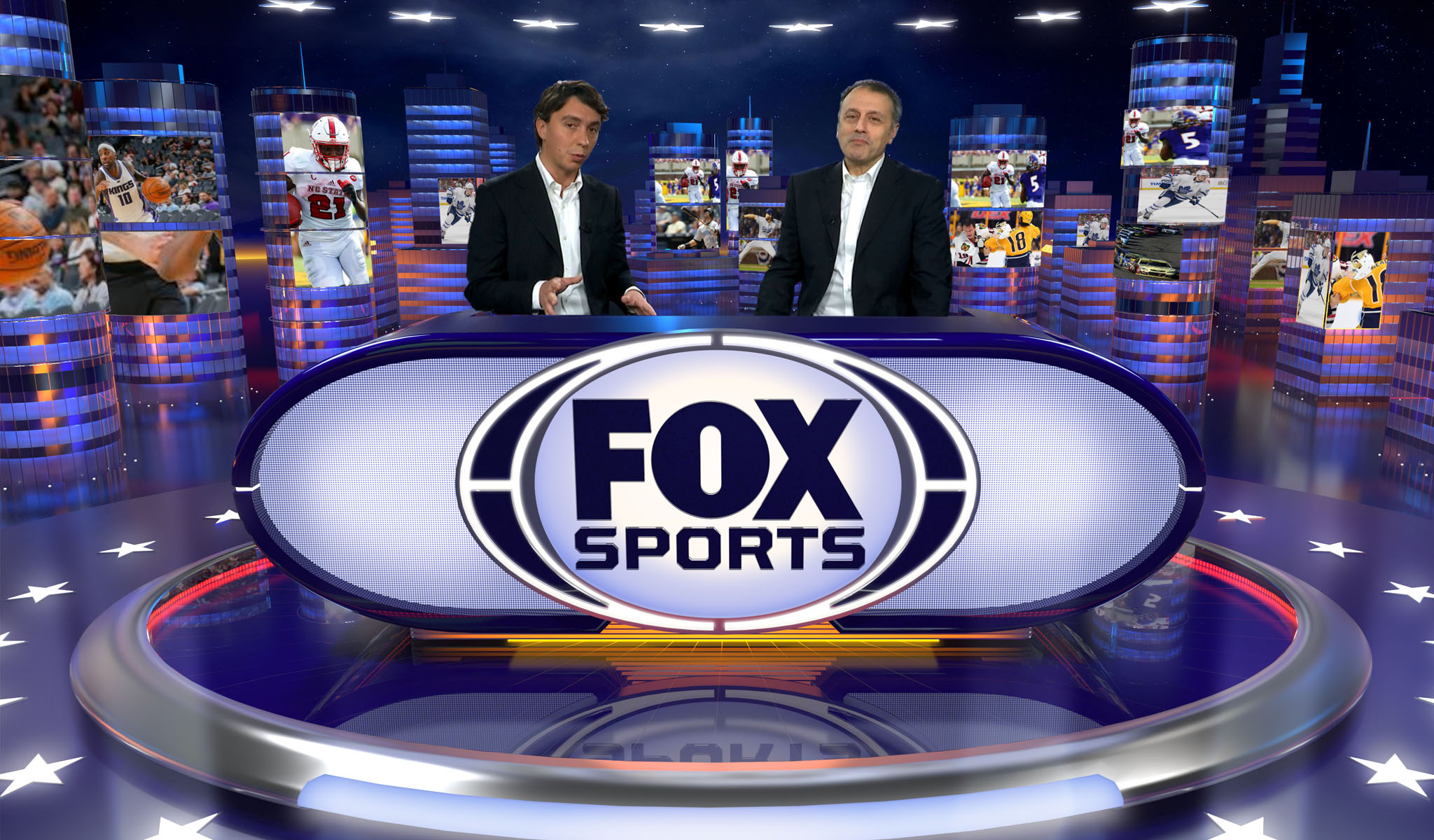 Two men in suits at a FOX Sports desk with sports images and cityscape graphics in the background.