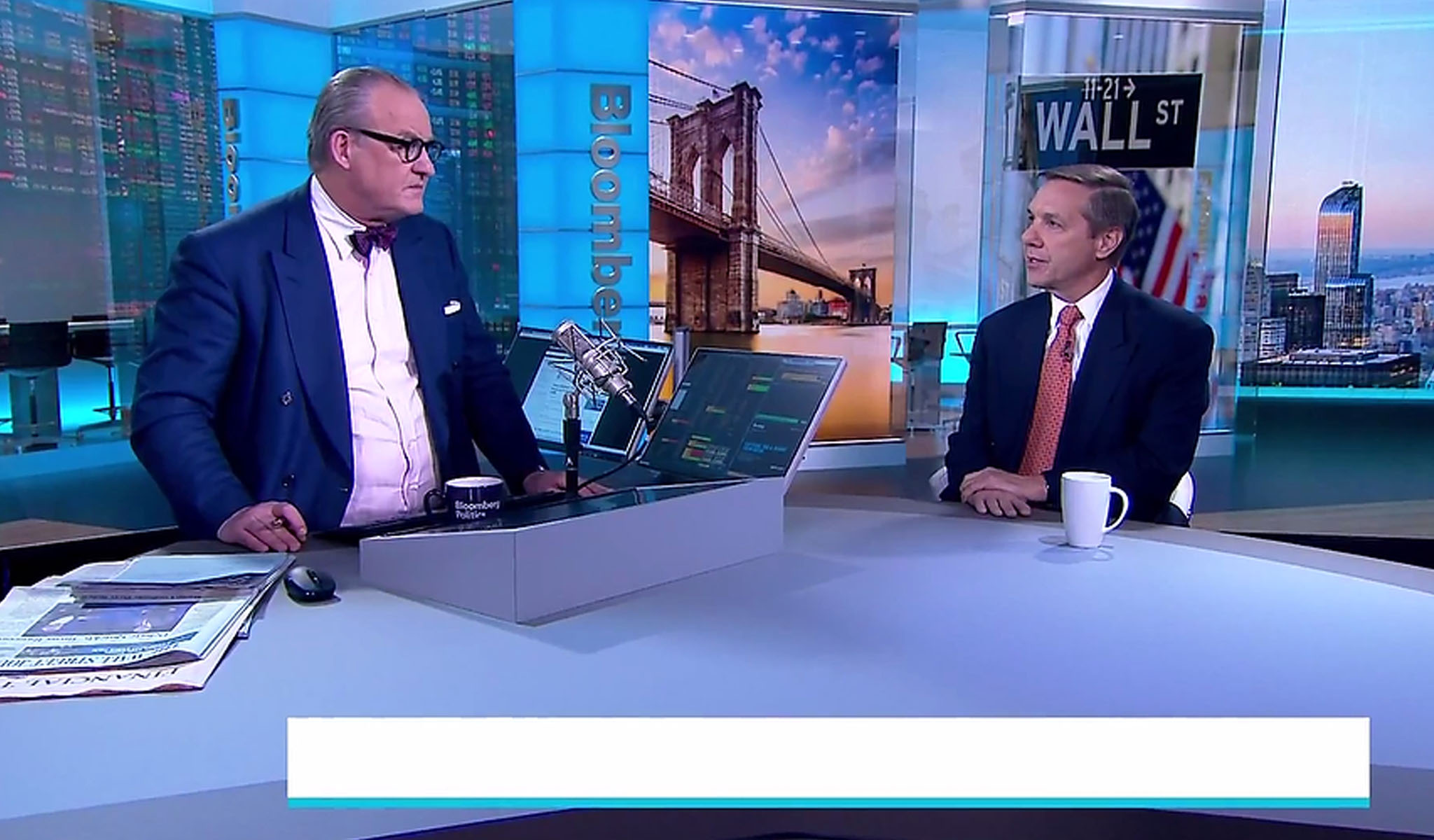 Two men in suits having a discussion in a Bloomberg TV studio with Wall Street and Brooklyn Bridge images.