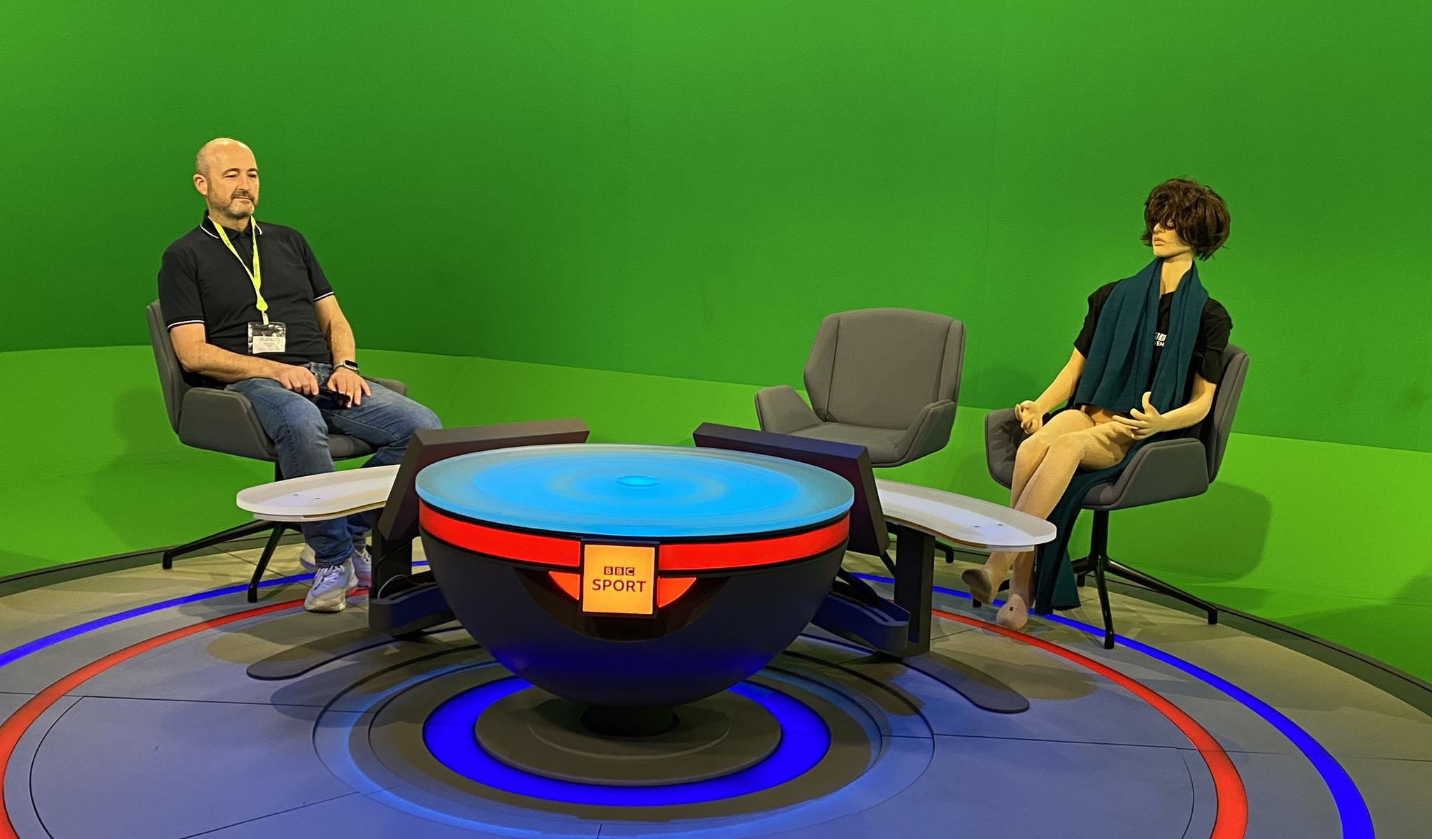 Man and mannequin seated in chairs around a BBC Sport studio set with green screen background.