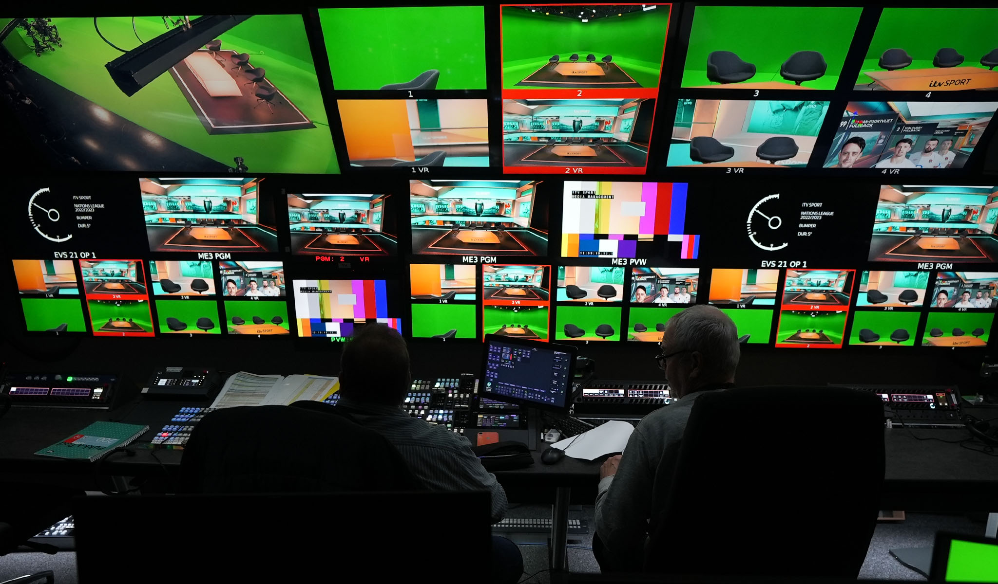 Two men working in a TV control room with multiple screens showing green screens and studio shots.