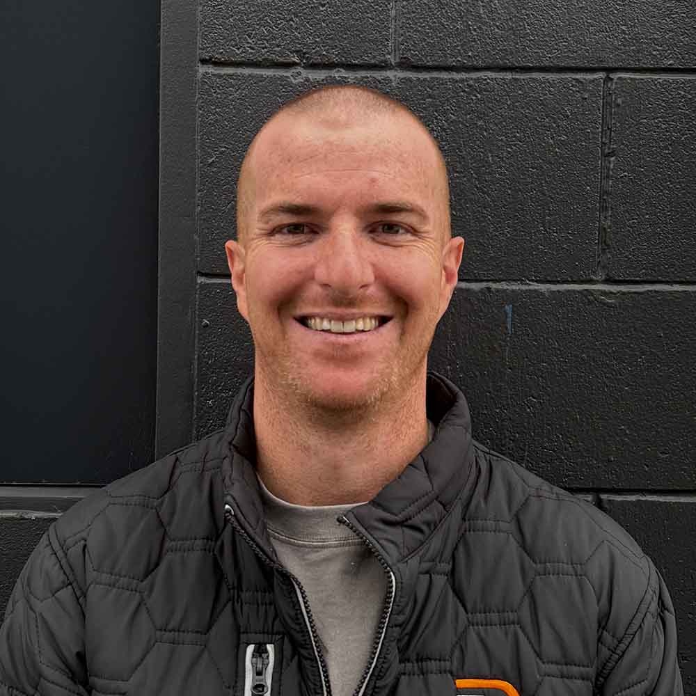 Smiling man with short hair wearing a black quilted jacket standing against a black brick wall.