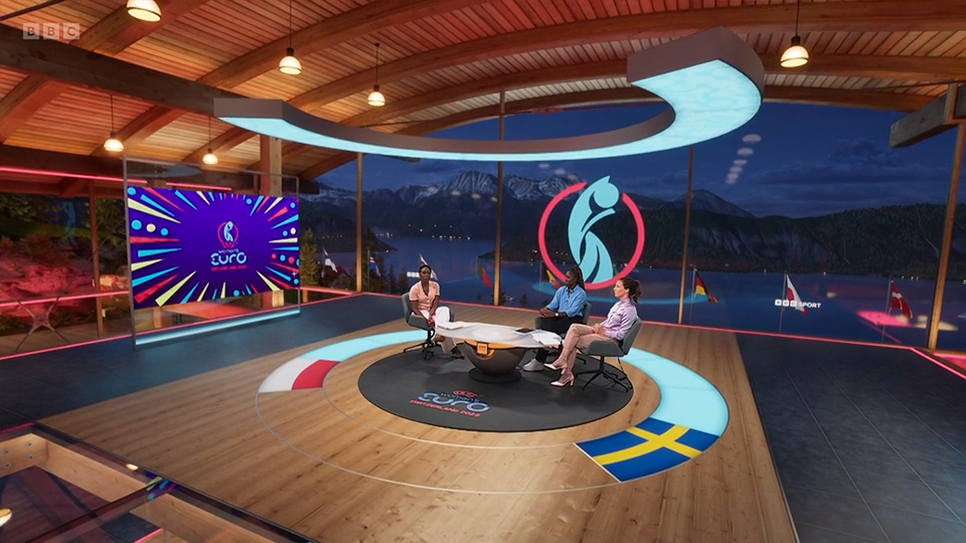 A high shot of Jeanette Kwakye and guests inside the BBC Sport virtual set for Sweden v Poland at Euro 2025. The set is a timber pavilion with a vaulted roof set above Lake Lucerne. In the background we can see mountains. Because it is night time, lights from houses and roads line the egde of the lake.