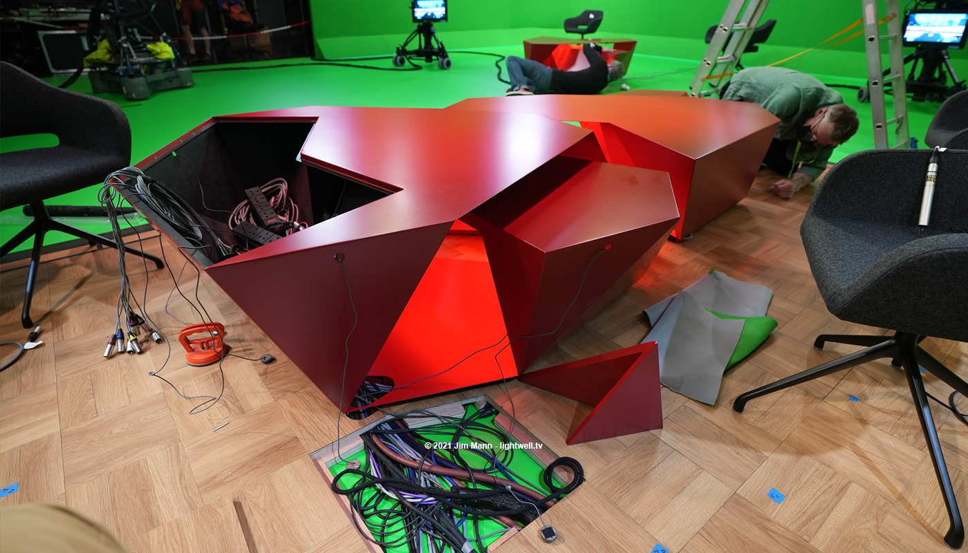 The presenters desk from BBC Tokyo 2020 is shown open, revealing cabling for microphones and prompter screens. Members of the design team and studio crew work in the background