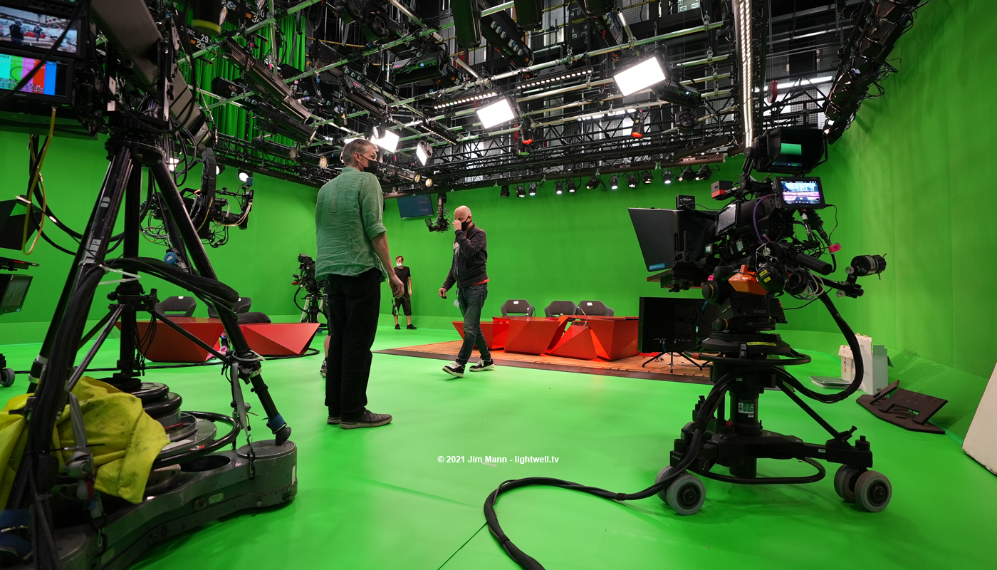 low angle view of the BBC Tokyo set being installed and rigged ahead of broadcast. The desk is partly assembled with elements propped against the greenscreen. Toby Kalitowski surveys the space, whilst lighting director Dave Gibson walks towards him ready to discuss the lighting