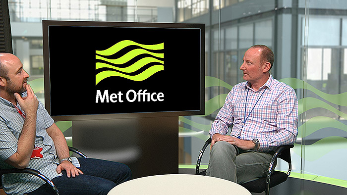 A presenter and guest sit in front of a screen displaying the Met Office logo. They are deep in conversation. The virtually re-constructed Met Office HQ interior is in the background.